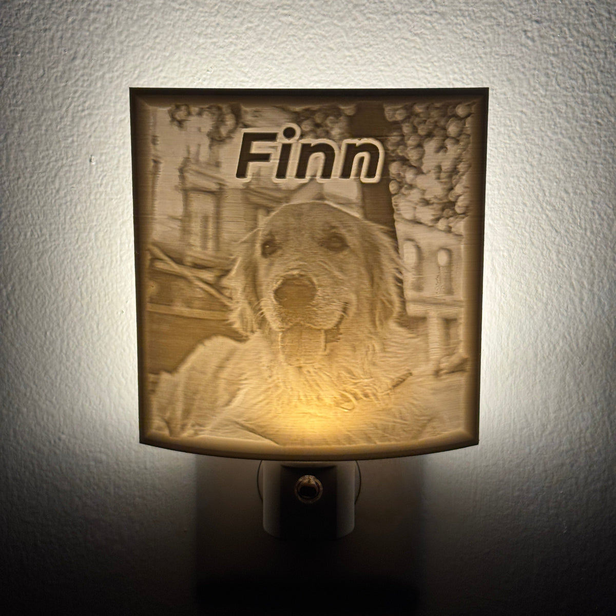 A personalized pet memorial night light glowing on a textured wall. The square light fixture features an engraved design of a golden retriever with the name 'Finn' written above it, casting a warm yellow glow.