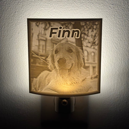 A personalized pet memorial night light glowing on a textured wall. The square light fixture features an engraved design of a golden retriever with the name 'Finn' written above it, casting a warm yellow glow.